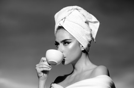 Close up portrait of woman holding a cup with coffee at sunrise. Beauty woman with towel on head drinking coffee in the sun, outdoor in sunlight light, enjoying morning coffee.の写真素材
