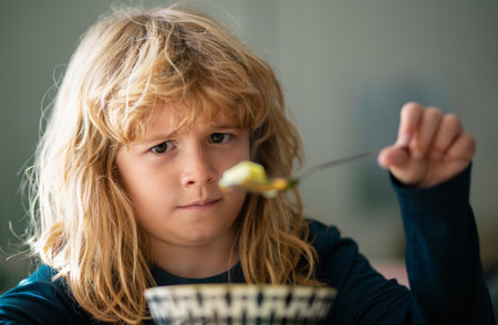 Close up portrait of funny kid eating. Sad boy eating healthy chicken noodle soup for lunch. Unhappy baby child taking food at home or nursery daycare or kindergarten.の写真素材