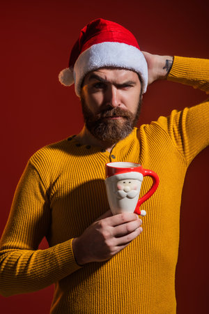 Santas face. Bearded man in Santa hats on red studio background. Santa celebration Happy New Year and Christmas winter holiday. Portrait of Santa with cup. Man beard in Santa costume with cup.の写真素材