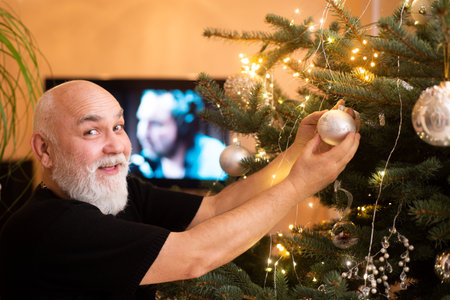 Happy Senior man Decorating Christmas Tree at Home. Senior Putting up Christmas Decorations during New Year Time at Home. A portrait of senior man with Christmas Bauble at Christmas time.の写真素材