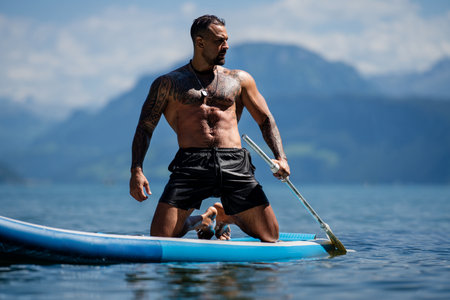 Man on paddle board in Alps lake mountains. Leisure activities with paddles on Lake. Man paddle surfing board. Muscular strong Hispanic man paddle board surfers. Latin man on sup board paddle surfing.の写真素材