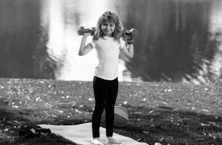 Sport child with strong biceps muscles. Kids exercising fitness dumbbells. Strong little boy exercising with dumbbell in park outdoor. Sport strong boy, little bodybuilder.の写真素材