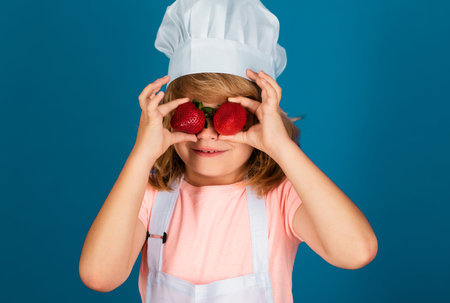 Child chef isolated on blue. Funny little kid chef cook wearing uniform cook cap and apron hold strawberries cooked food in the kitchen.の写真素材