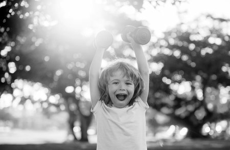 Fitness dumbbells, excited kid exercise workout outdoor. Amazed boy, sporty child with dumbbells in summer park.の写真素材