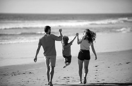 Back view of happy young family walking on beach. Child with parents holding hands. Full length poeple.の写真素材