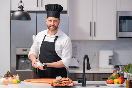 Millennial hispanic man in chef apron and cook hat hold salmon fillet at kitchen. Cooking salmon fillet. Restaurant menu with salmon fillet. Cooking, advertising salmon fillet concept.の写真素材