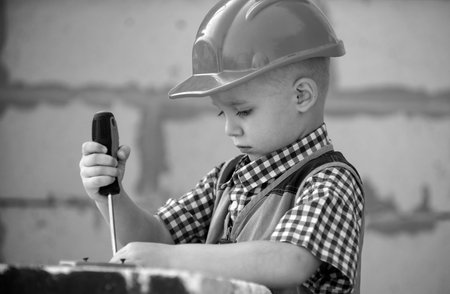 Kid boy twists bolt with screwdriver. Child repairman with repair tool. Child in helmet and boilersuit on construction site. Little worker engineer. Repair home.の写真素材