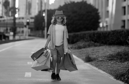 Cute little boy in summer fashion clothes goes shopping. Happy child with shopping packages in hands. Shopper child with shopping bag outdoor. Cute kid walking on street carrying shopping bags.の写真素材