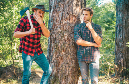Woodcutters lumberjacks. Hipsters men on serious face with axe. Lumberjack brutal and bearded holds axe. Two lumberjacks in forest. Man in hat looks brutally.の写真素材