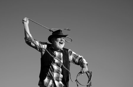 Senior western cowboy throwing lasso rope. Bearded wild west man with brown jacket and hat catching horse or cow. Rodeo or ranch.の写真素材