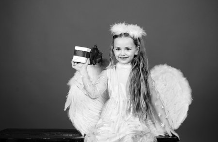 Kid girl angel with present gift, studio portrait. Little angel with white wings holds gift, happy fairy. Cute angel child girl with angels wings, isolated on red. Valentines day.の写真素材