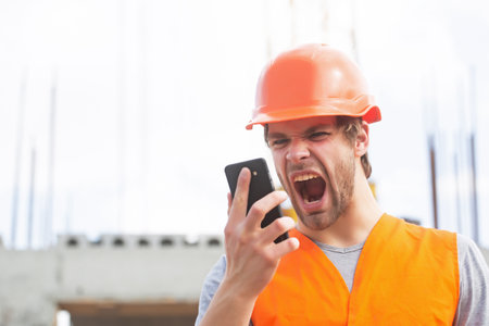 Man builder worker in helmet screaming in mobile phone on construction site. Portrait of man builder...
