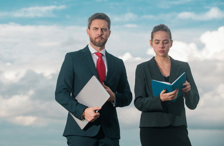Successful business couple. Businessman and businesswomen. Successful business people standing over blue sky. Man hold laptop woman with notebook.の写真素材