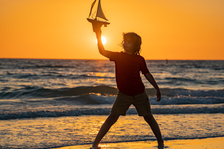 Dream of summer. Little blonde boy put toy boat in the sea waves at the beach. Summer with kids vacation. Dream on travel. Silhouette of kid playing with toy seailing boat on sunset sea.の写真素材