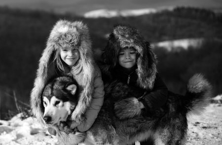Funny little children embraced husky dog in winter outdoor. Smiling kids friends hug dog in frost snowy day outdoor. Two handsome boys an girl resting together in park with snow background.の写真素材