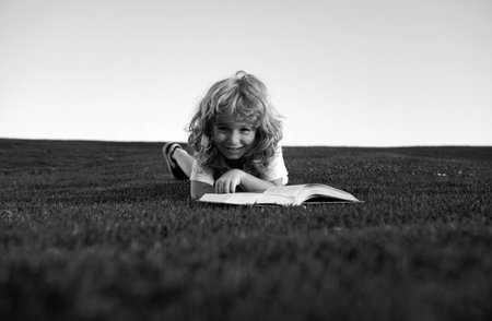 Smart kid boy reading book in park on green grass lawn outdoor.