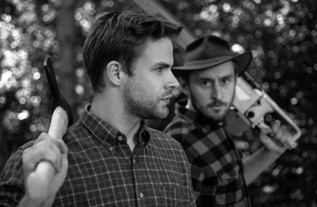 Lumberjack workers in the forest. Woodcutter with axe and lumberjack with chainsaw. Handsome...