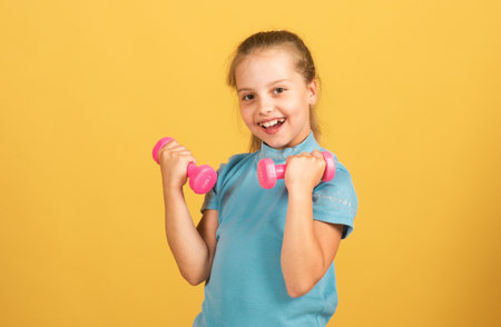 Cute little girl doing exercises with dumbbells in yellow background. Closeup portrait of sporty child with dumbbells. Happy child girl exercising.の写真素材