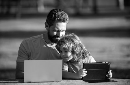Father teaching son to use laptop, dad and school boy child looking computer screen and tablet, playing game, watching video, sitting on grass. Outdoor family weekend.の写真素材
