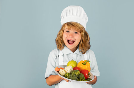 Excited funny chef cook. Portrait of a 7, 8 years old child in cook cap and apron making fruit salad and cooking food in kitchen. Cute little blonde happy smiling chef hold plate with vegetables.の写真素材