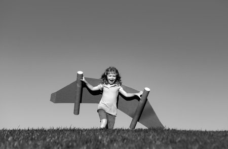 Happy child playing outside on green grass and blue sky. Kid pilot with toy jetpack. Kid boy play with toy plane cardboard. Summer travel and adventure. Success winner, imagination and dream concept.の写真素材