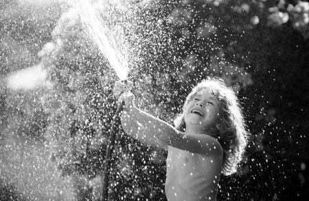 Cute little boy watering flowers in the garden at summer day. Child farmer with garden hose in planting plant. hose. Funny kid watering plants in the yard garden.の写真素材
