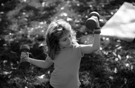 Boy is doing exercises to develop muscles. Child is doing exercises with dumbbells in park.