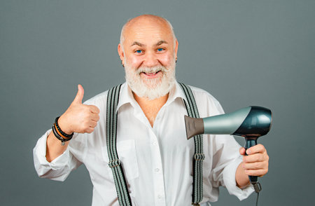 Senior hairdresser or barber with happy emotional face in studio on grey background. Man with Blow-dry with thumb up.の写真素材