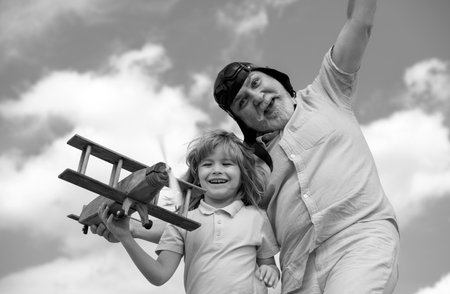 Funny grandson child and grandfather playing with wooden plane against summer sky background. Child boy with dreams of flying or traveling.の写真素材