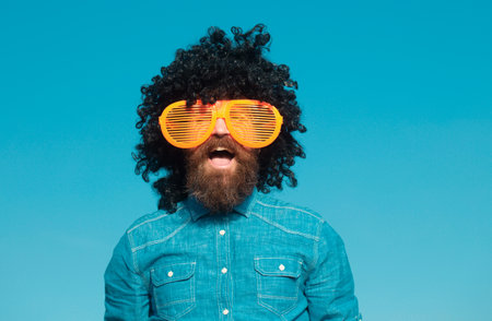 Crazy funny bearded man with wig and fun glasses on sky background. Funny bearded man in wig. Handsome bearded man with mustache in wig.の写真素材