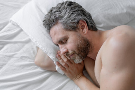 Male head on pillow. Gray hair man sleep in white bed. Senior man sleep in bed at bedroom. Mature man sleeps at home at morning. Good sleep. Millennial man sleeping in bed. People bedtime, rest sleep.の写真素材