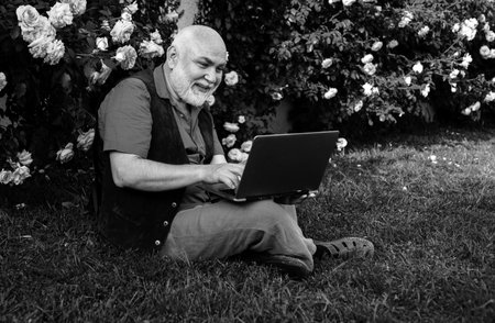 Telework concept. Senior man in garden with laptop. Old gardener online social network games.の写真素材