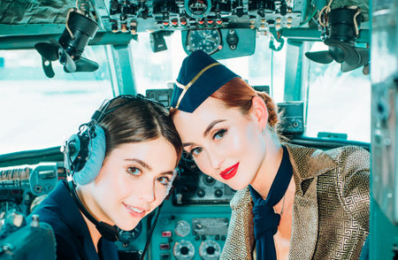 Portrait of two smiling women pilots. Beautiful Smiling Young Woman Pilot Sitting in Cabin of Modern Aircraft. Stewardess and Flight Instructor in an Aircraft Cockpit. Pilot and Stewardess.の写真素材