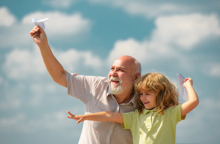 Child boy and grandfather with paper plane over blue sky and clouds. Men generation granddad and grandchild. Elderly old relative with child.の写真素材