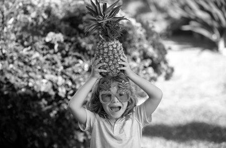 Funny child holds a pineapple on her head on a summer nature background. Pineapple kid boy and pineapples on summer vacation.の写真素材