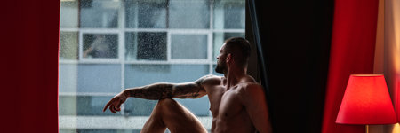 Man window on rainy weather looking through the window with rain drops, autumn banner. Handsome muscular man in bedroom on window curtains. Young handsome sexy man resting at home.の写真素材