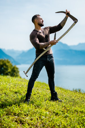 Strong farmer using scythe. Sexy Farmer with a scythe on green grass field. Muscular Farmer cut grass. Rural farmer on summer meadow mowing grass with classic traditional scythe.の写真素材