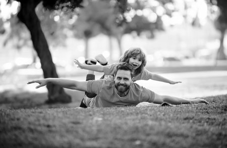 Father lying on grass, with excited happy little child son on shoulder. Carefree two man generations family having fun. Weekend man family concept.の写真素材
