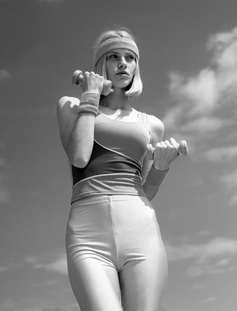 Blonde girl doing sport exercises with dumbbells at sky background. Leisure time sport activities. Beautiful girl take care about her health drinking water and doing sport exercises.の写真素材