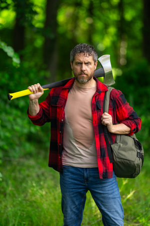 Confident lumberjack works in summer forest. Man with an ax. Woodcutter in plaid shirt in forest. Hipster with axe. Lumberjack with an axe. Bearded lumberjack. American lumberjack with hatchet.の写真素材