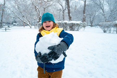 Happy Kid in winter park on a snowy cold winter day. Cute child playing with snow in winter. Portrait of funny child play with snowman. Winter activities Christmas. Kid building snowman in park.の写真素材