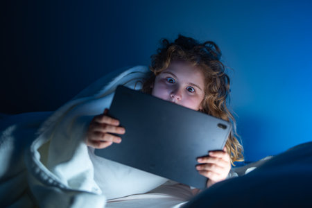 Kid lying on bed and surfing Internet on tablet in dark room. Boy reading Tablet in bed at night....