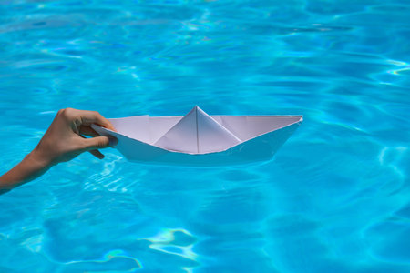 Female hand holding paper boat on the sea background. Origami paper boat sailing on water causing waves and ripples.の写真素材