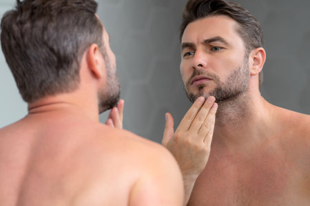 Sexy sensual man with perfect skin touch face after shaving. Skin care healthcare cosmetic procedures concept. Close up man looking in mirror, sensitive skin, cosmetology treatment. Skin care.の写真素材