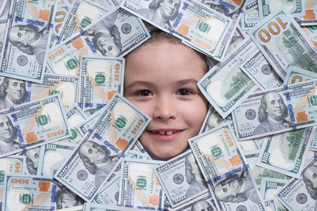 Kid with fun face in money. Kid peeking out of dollar bills with astonished shocked eyes. Money background.の写真素材