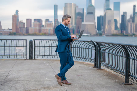 Mature hispanic businessman at the city. Businessman with laptop on streets of New York Citys financial district. Businessman at Manhattan. Businessman works on laptop outdoor. Business in NYC.の写真素材