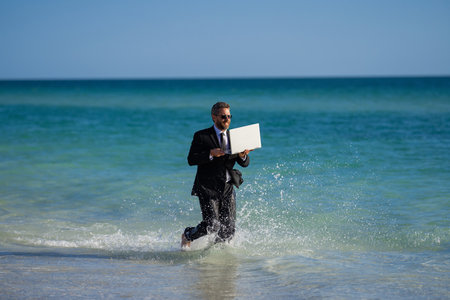 Leisure weekend and remote freelance work. Crazy comic business man in suit rest on sea bech. Funny businessman with laptop runnig on water. Remote summer business. Summer holiday for businessman.の写真素材