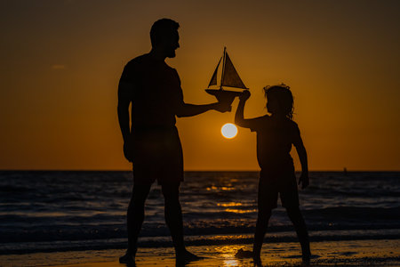Parenting, fatherhood and fathers day concept. Silhouttes of father with son on seaside at sunset. Fathers day. Father and son dream on travel. Father and son play with toys boat. Summer travel.の写真素材