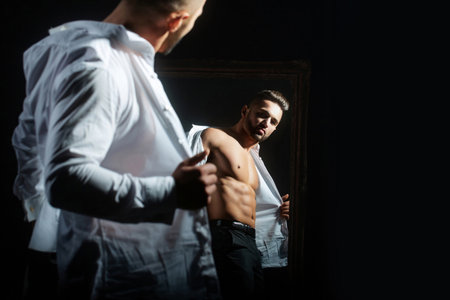 Fashion male model with hot naked body in white shirt. A sexy man in an unbuttoned white shirt on black background. Handsome muscular man with naked torso and shirt. Guy with with open shirt.の写真素材