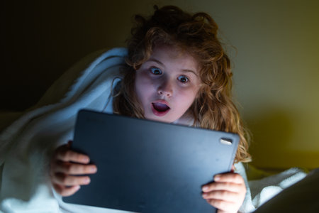 Child using Tablet in bed at night. Kid in pajamas using tablet before going to bed. Kid using gadgets. Child watching his tablet in bed. Kid lying on bed and surfing Internet on tablet in dark room.の写真素材